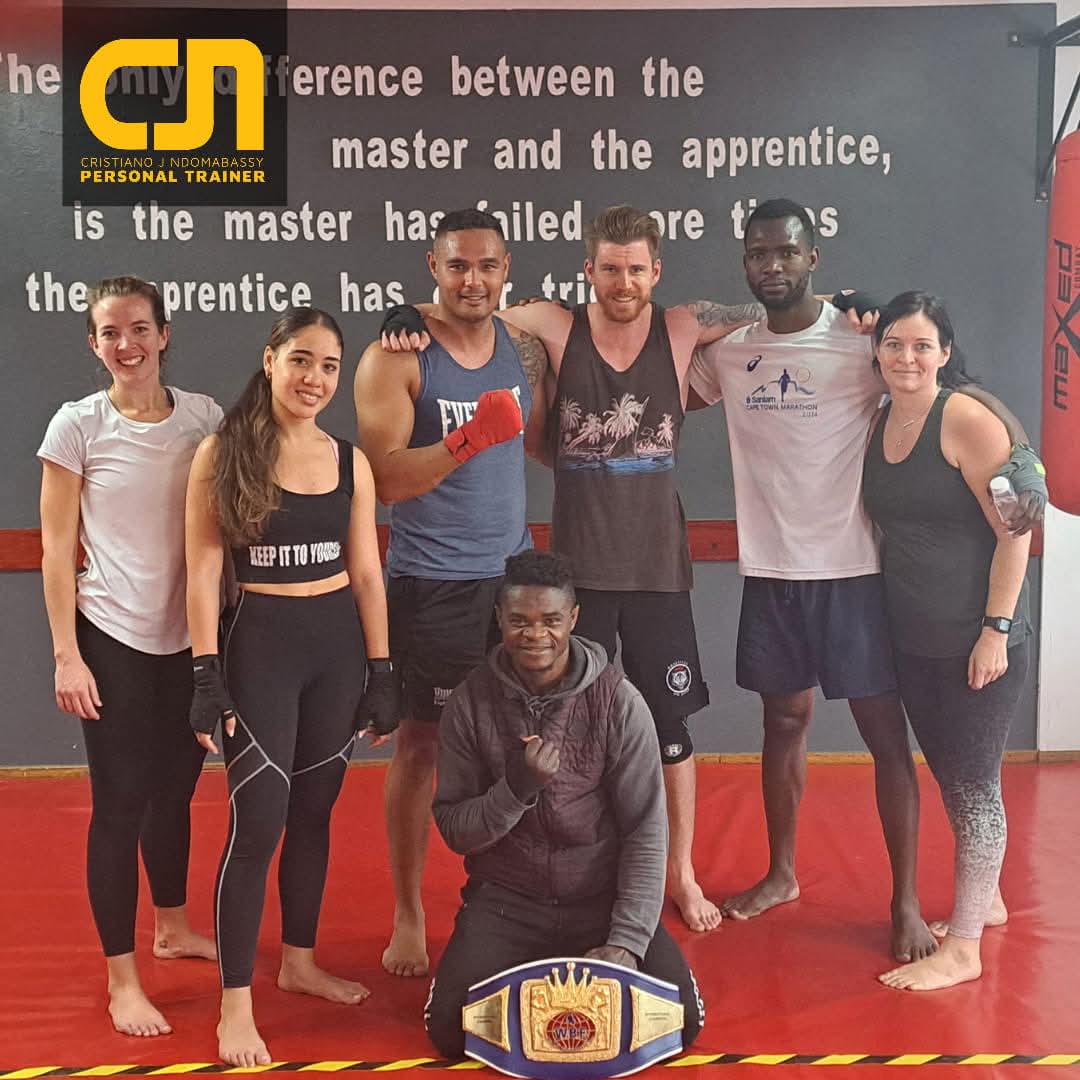Group of seven people standing and kneeling on a red mat in a martial arts gym, some wearing boxing gloves and athletic clothing, one person holding a championship belt. The mood is friendly and celebratory.